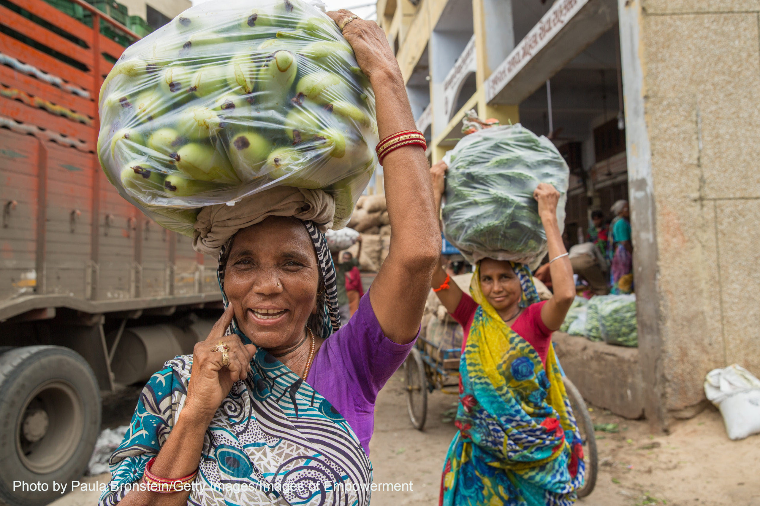 two women smiling carrying vegetables on their heads in front of a truck